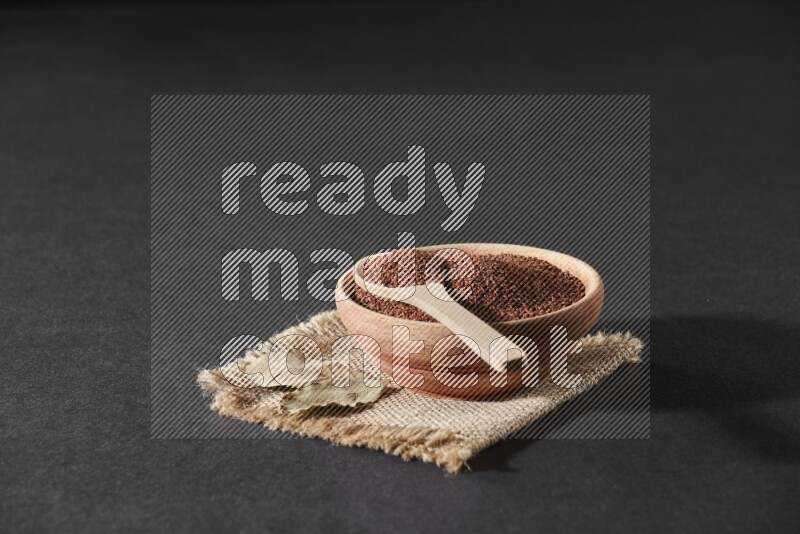 A wooden bowl full of garden cress seeds with wooden spoon full of the seeds on it on burlap fabric on a black flooring