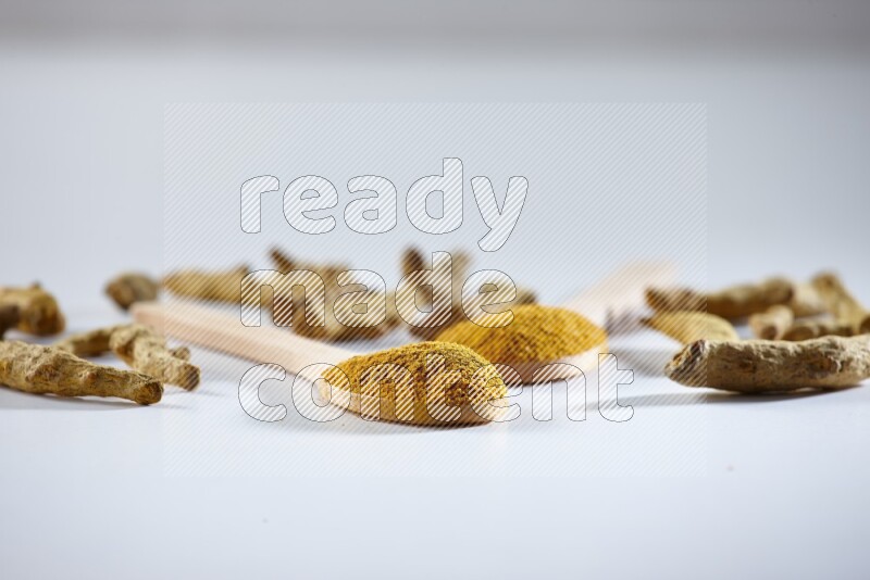 ملعقتان خشبيتان ممتلئتان ببودرة الكركم مع أصابع الكركم المجففة بجانبهما على خلفية بيضاء