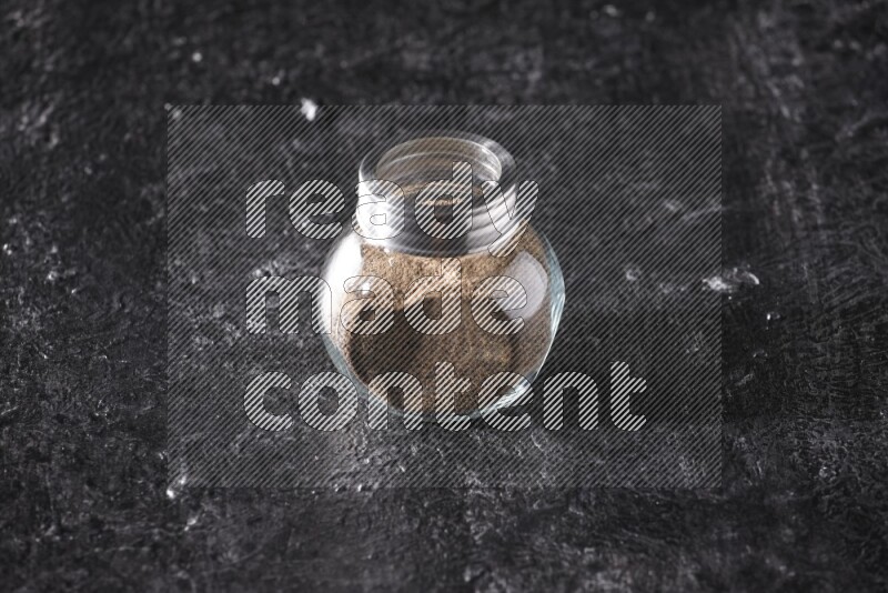 A glass spice jar full of black pepper powder on textured black flooring