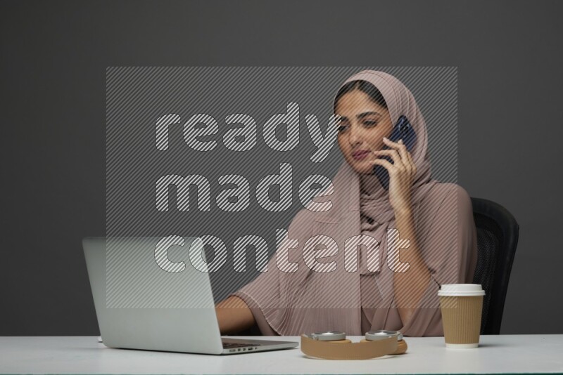 A Saudi woman Setting on her desk
 calling  on a Gray Background wearing Brown Abaya with Hijab