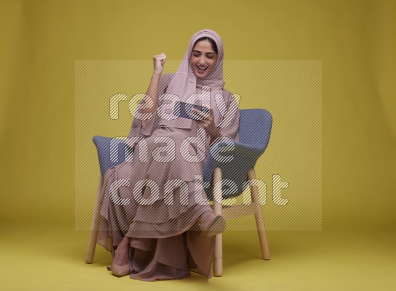 A woman Playing Game Sitting  on a Yellow Background wearing Brown Abaya with Hijab