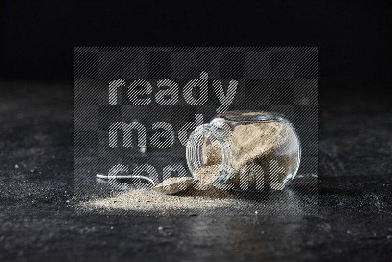 A flipped herbal glass jar and a metal spoon full of white pepper powder with spilled powder on textured black flooring
