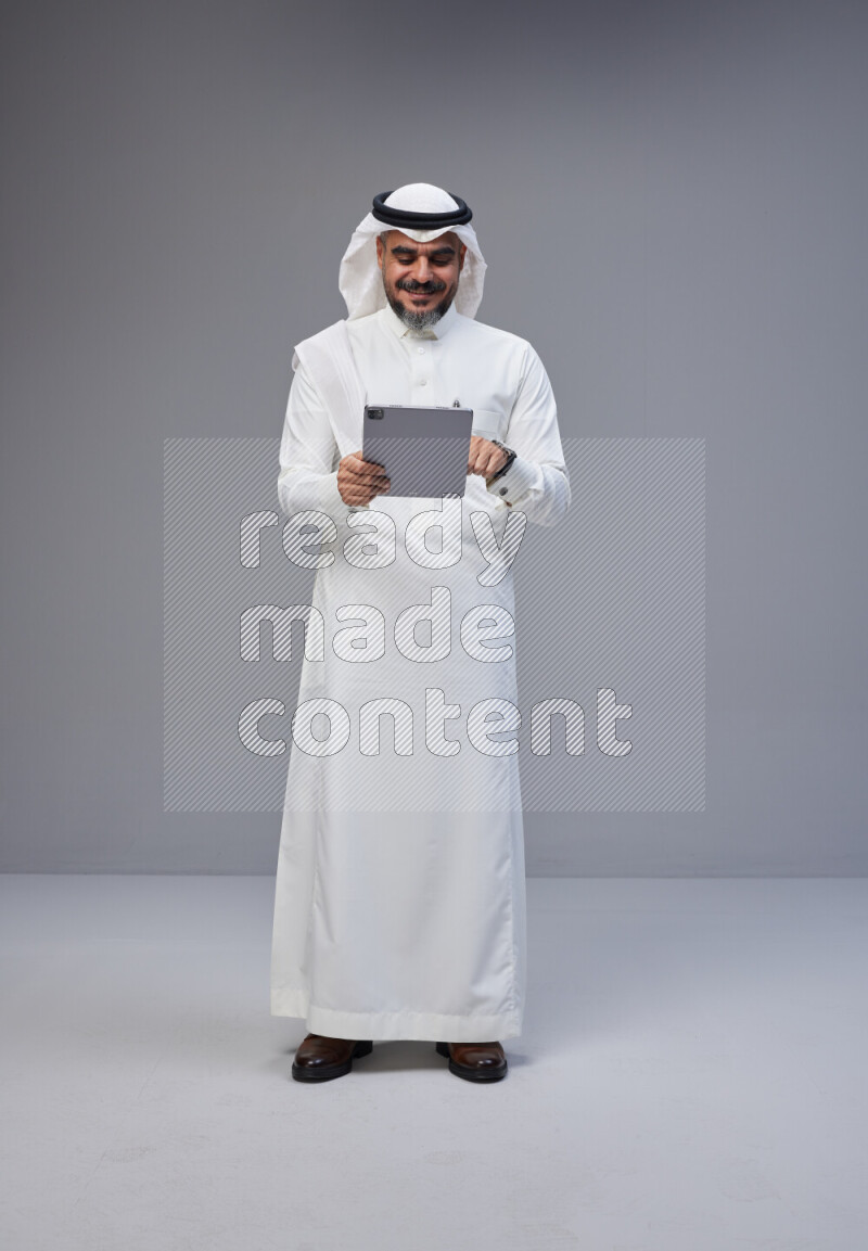 Saudi man Wearing Thob and white Shomag standing working on tablet on Gray background