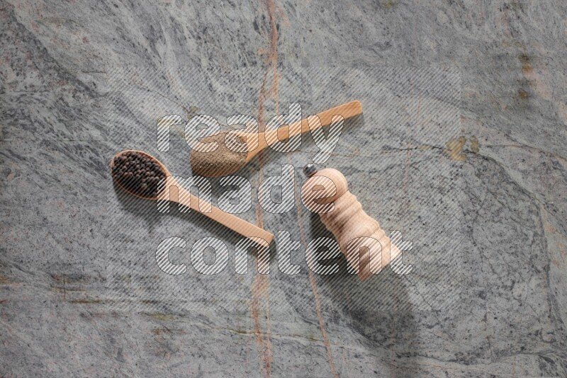 ملعقتان خشبيتان، ملعقة ممتلئة بمسحوق الفلفل الأسود والأخرى ممتلئة بحبات الفلفل الأسود ومطحنة خشبية على خلفية رخام رمادية