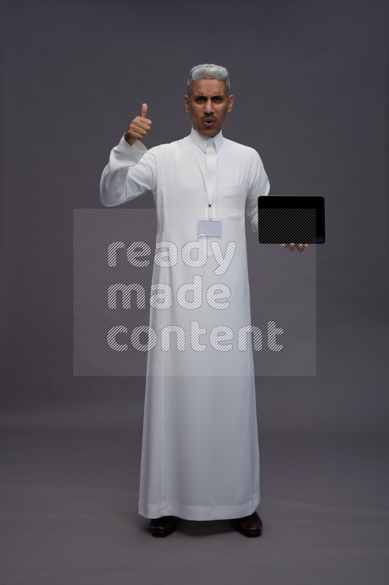 Saudi man wearing thob with neck strap employee badge standing showing tablet to camera on gray background