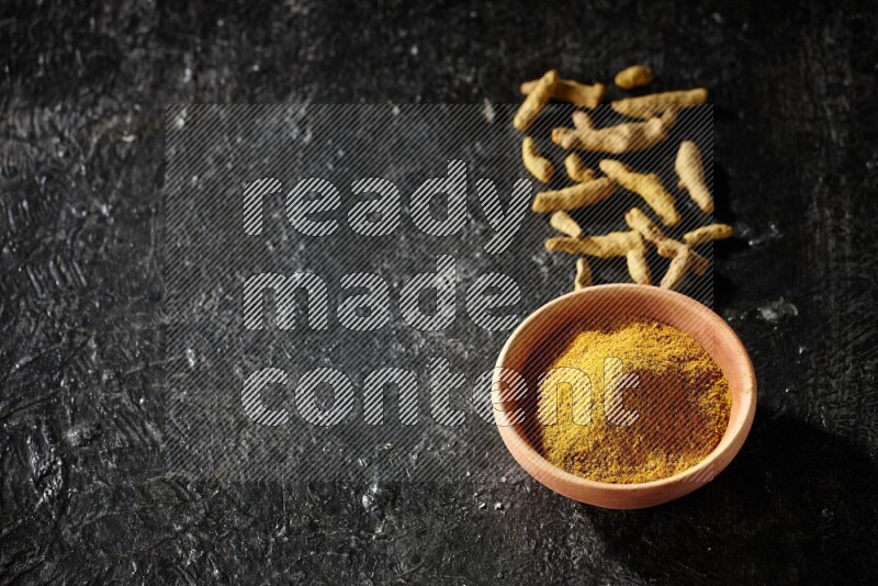 A wooden bowl full of turmeric powder with dried turmeric fingers on textured black flooring