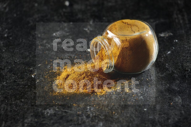 A flipped glass spice jar full of turmeric powder and powder spilled out of it on textured black flooring