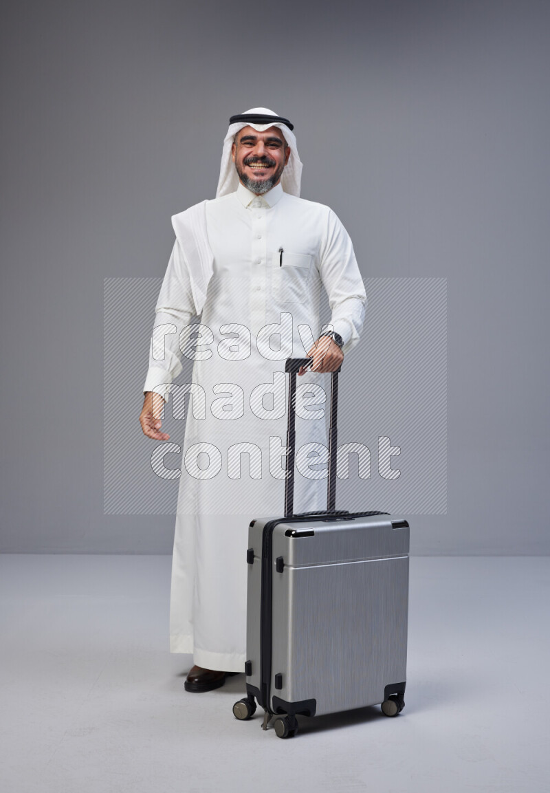 Saudi man wearing Thob and white Shomag standing holding Travel bag on Gray background