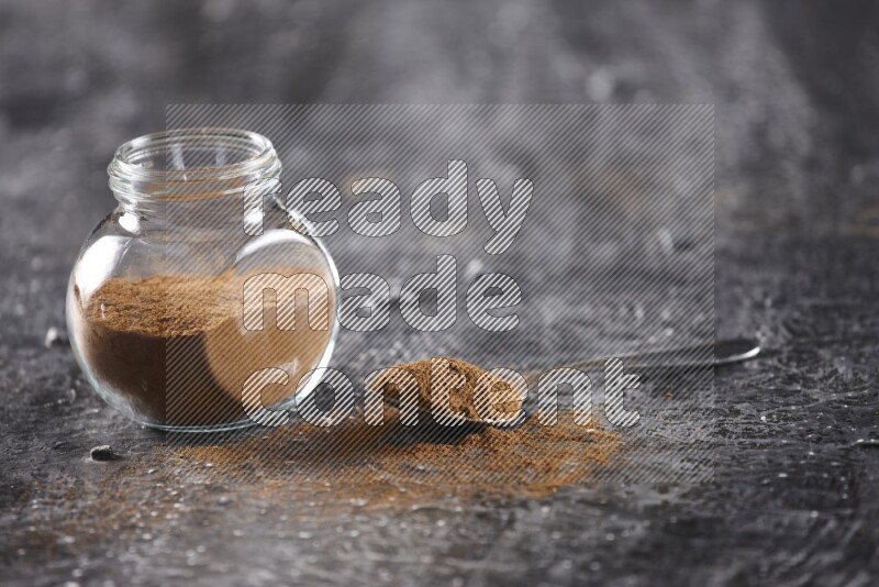 Herbal glass jar full of cinnamon powder and a metal spoon full of powder on textured black background
