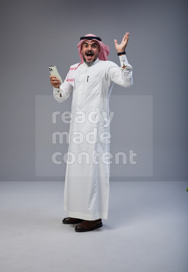 Saudi man Wearing Thob and red Shomag standing texting on phone on Gray background