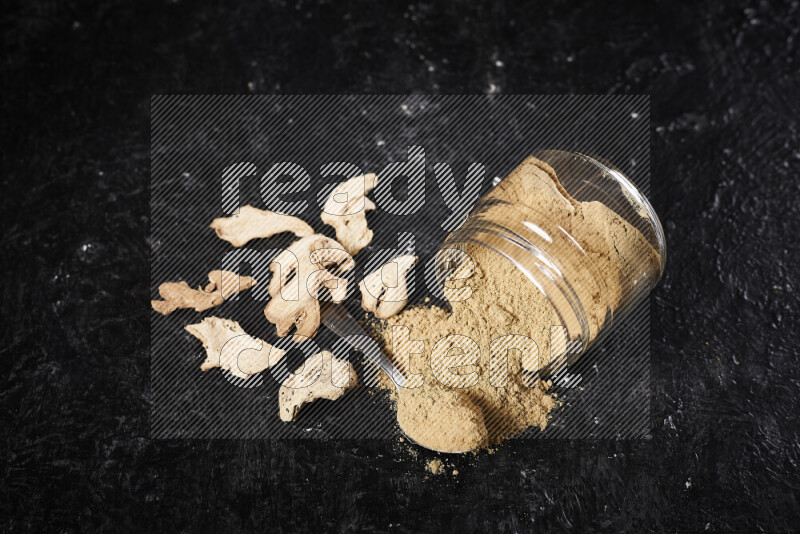 A glass jar full of ground ginger powder flipped with some spilling powder on black background