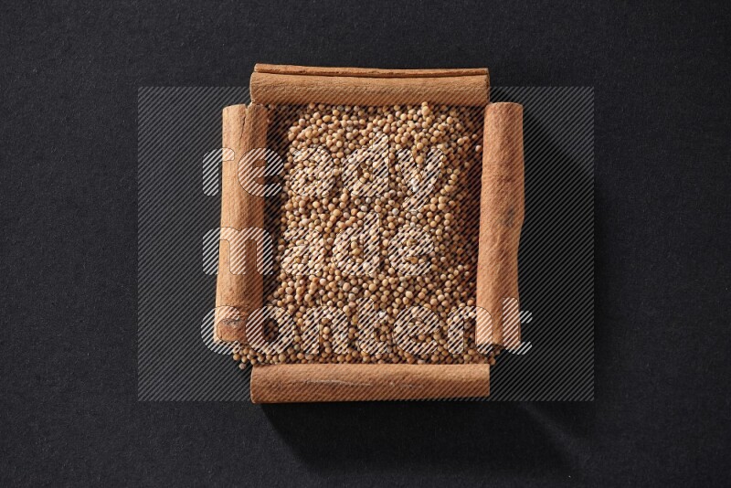 أربعة من أعواد القرفة مكونة شكل مربع بداخلها حبوب الخردل علي خلفية سوداء