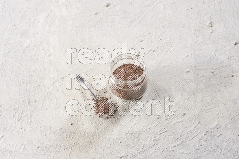 A glass jar full of flax seeds with a metal spoon full of the seeds on a textured white flooring