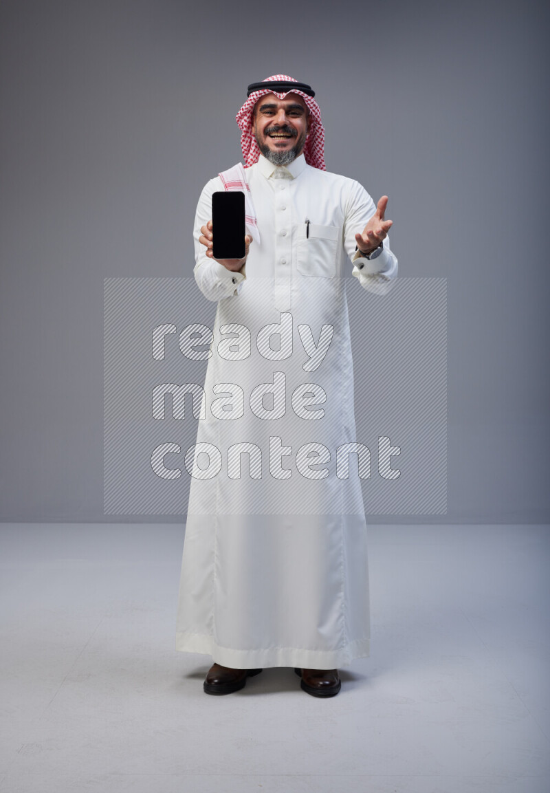 Saudi man Wearing Thob and red Shomag standing showing phone to camera on Gray background