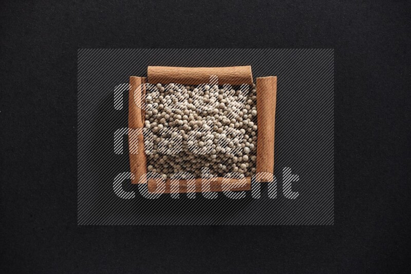 أربعة من أعواد القرفة مكونة شكل مربع بداخلها حبوب الفلفل الأبيض علي خلفية سوداء