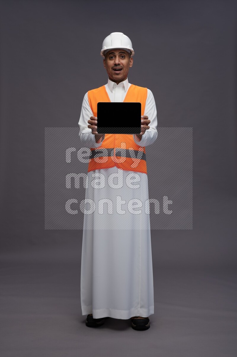 Saudi man wearing thob with engineer vest standing showing tablet to camera on gray background