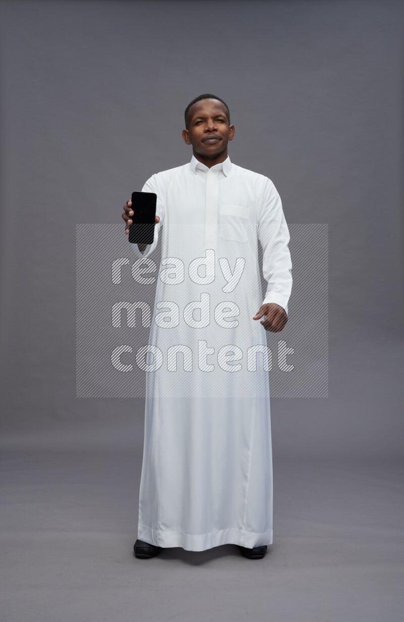 Saudi man wearing thob standing showing phone to camera on gray background