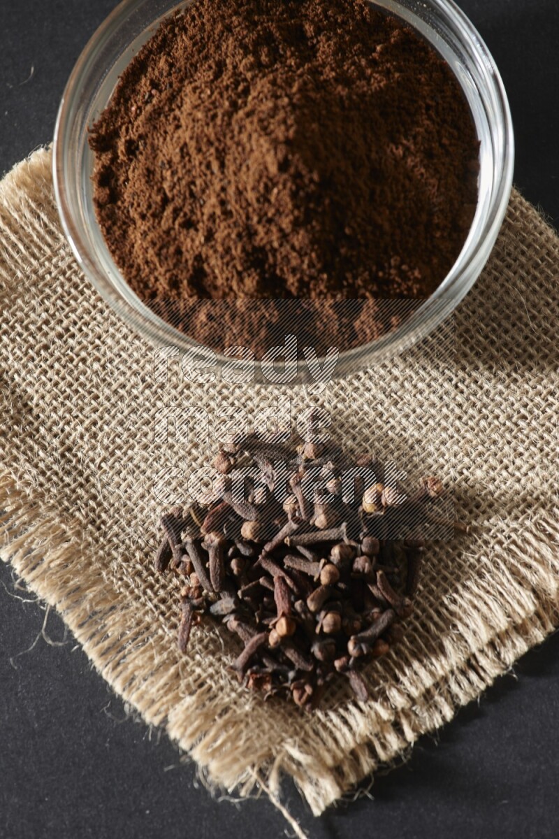 A glass bowl full of cloves powder with cloves grains on a burlap piece on a black flooring