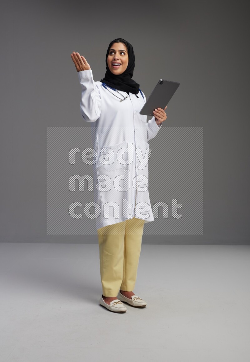 Saudi woman wearing lab coat with stethoscope standing working on tablet on Gray background