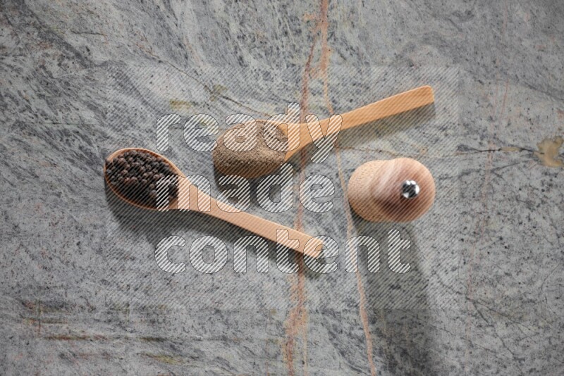 2 wooden spoons, one full of black pepper powder and the other full of black pepper beads and wooden grinder on a marble flooring