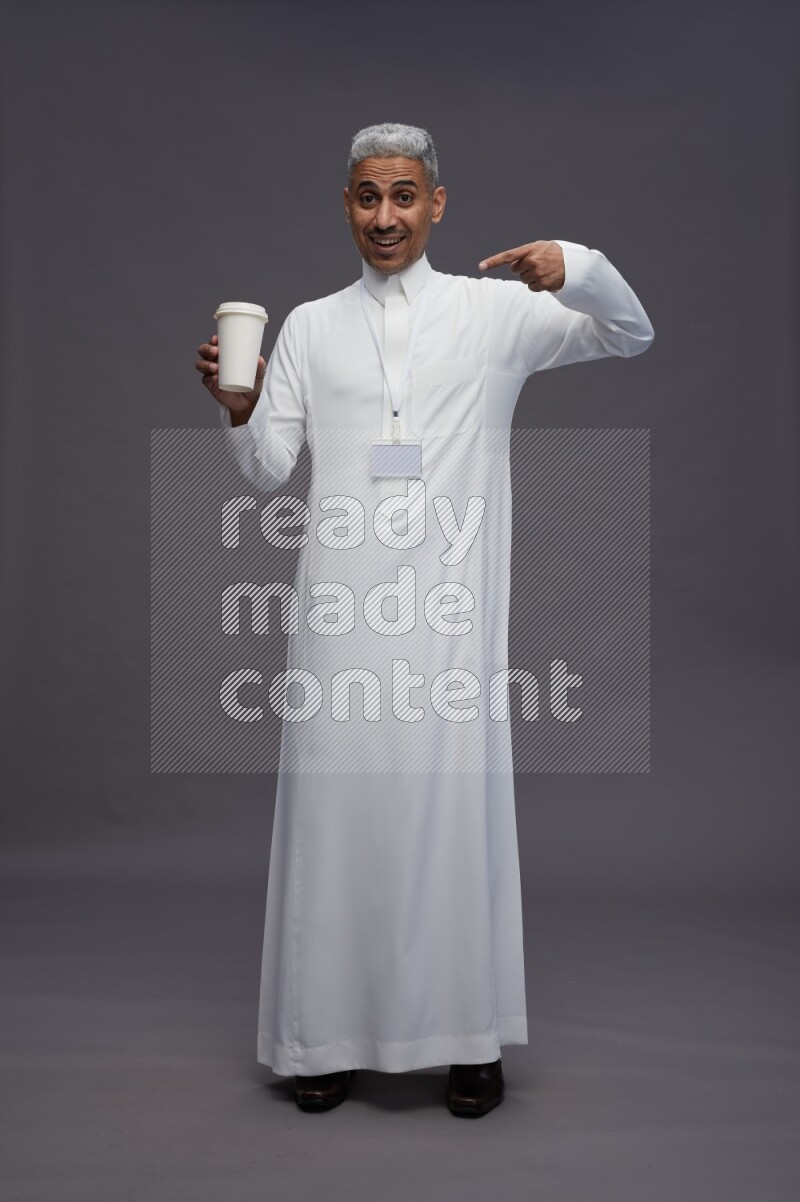 Saudi man wearing thob with neck strap employee badge standing holding paper cup on gray background