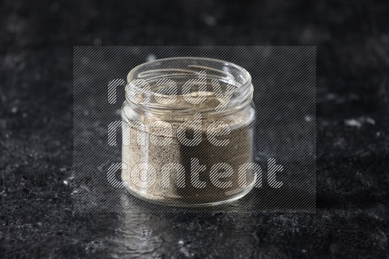 A glass jar full of white pepper powder on textured black flooring