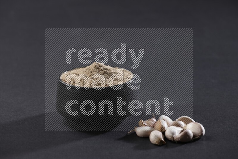 A black pottery bowl full of garlic powder with some cloves beside it on a black flooring