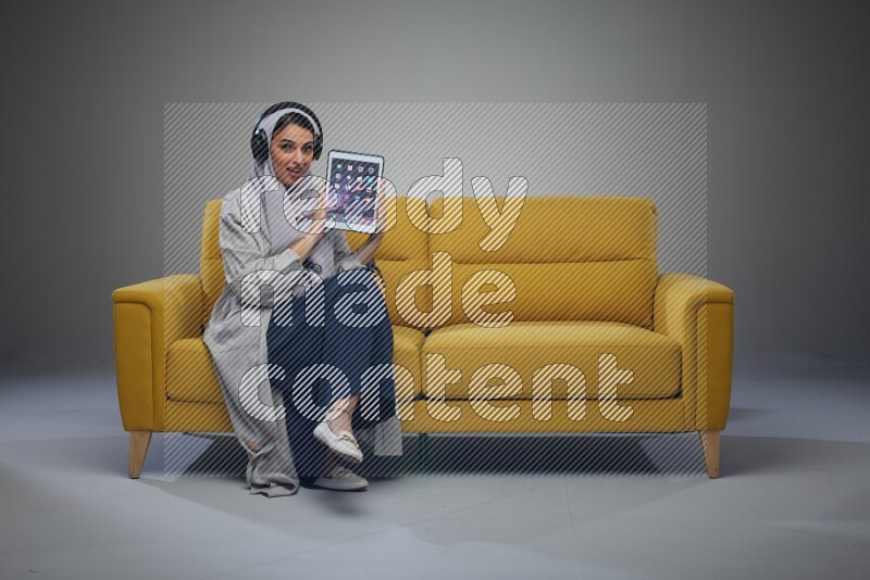 A Saudi female wearing a light gray Abaya and head scarf sitting on a yellow sofa and showing her tablet's screen while wearing headphones eye level on a grey background