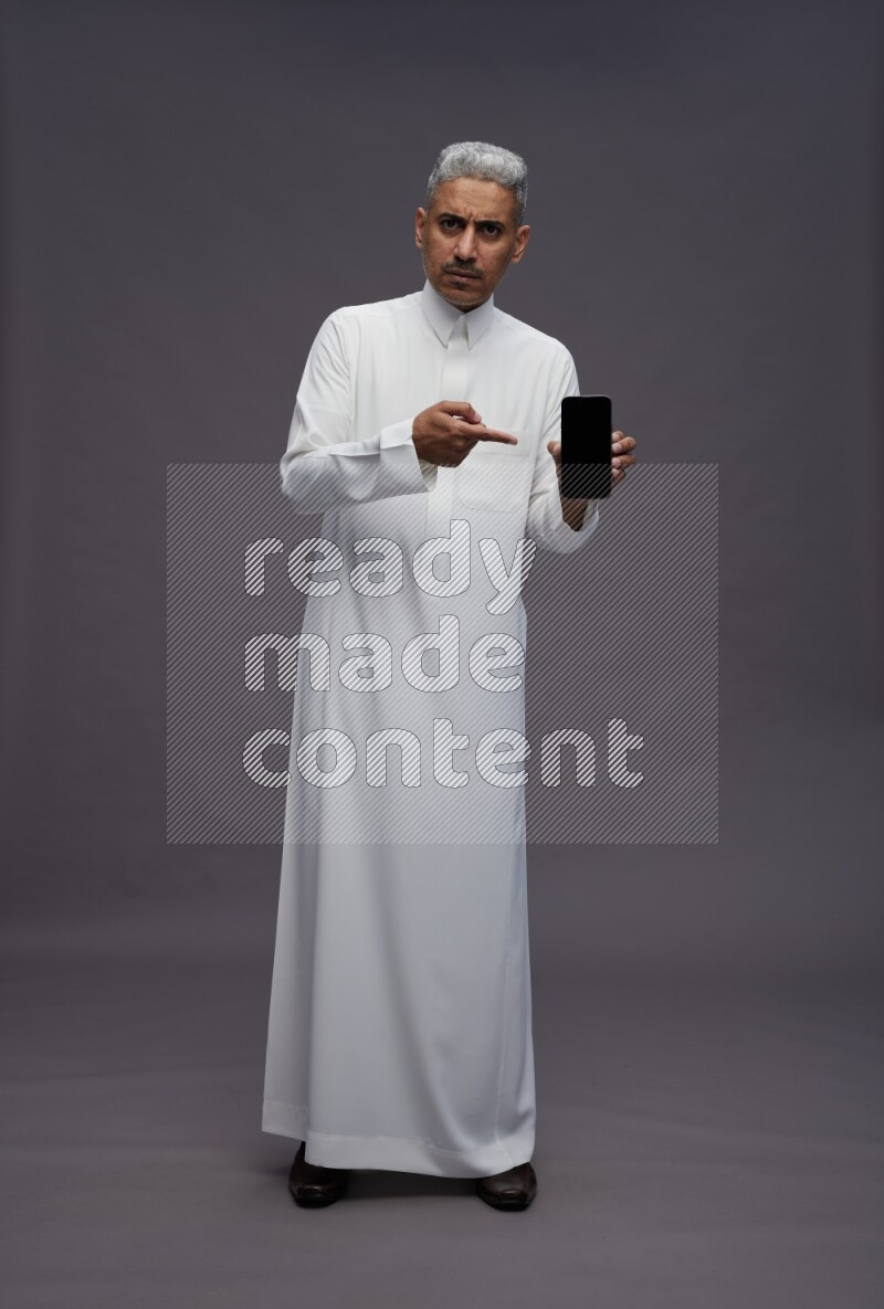 Saudi man wearing thob standing showing phone to camera on gray background