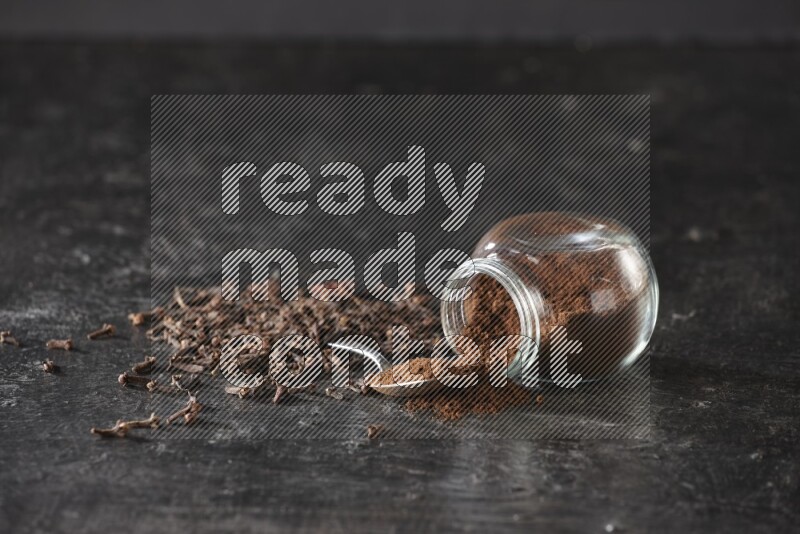 A flipped glass spice jar and a metal spoon full of cloves powder with cloves spread on textured black flooring