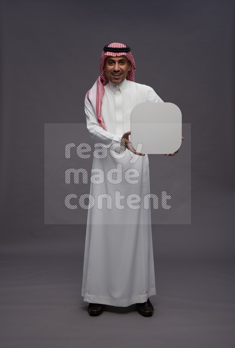 Saudi man wearing thob and shomag standing holding social media sign on gray background