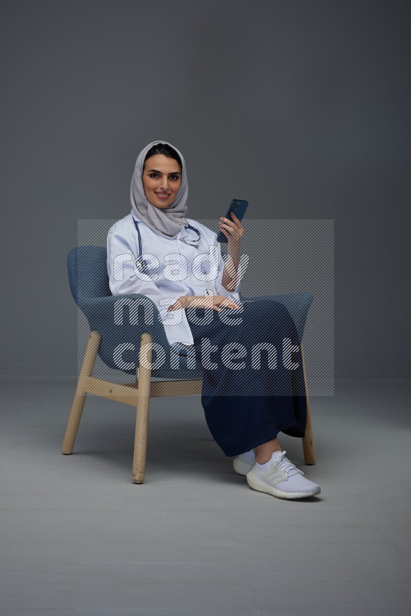 A doctor wearing a light gray head scarf sitting on a dark grey chair and using a laptop while talking in the phone eye level on a grey background