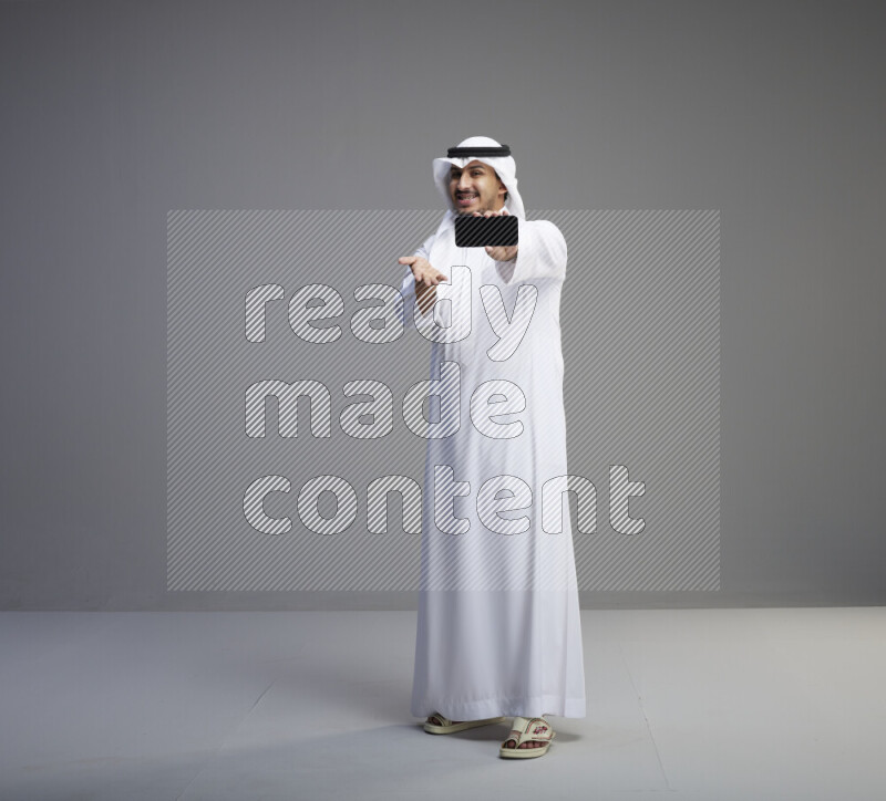 A Saudi man standing wearing thob and white shomag showing phone to camera on gray background