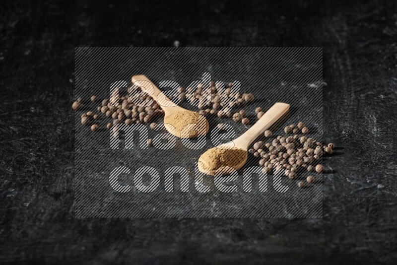 Wooden spoons full of all spice powder and allspice whole balls beside it on a textured black flooring