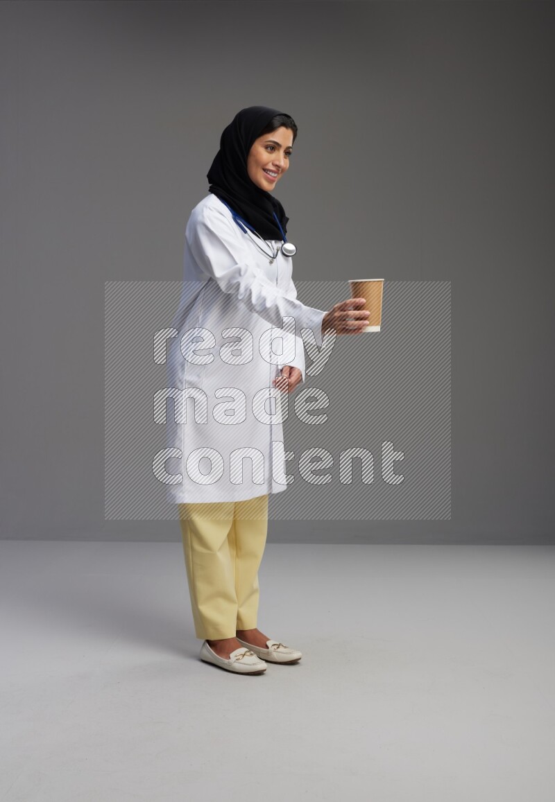 Saudi woman wearing lab coat with stethoscope standing holding paper cup on Gray background