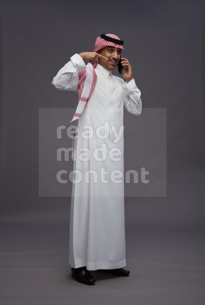 Saudi man wearing thob and shomag standing talking on phone on gray background