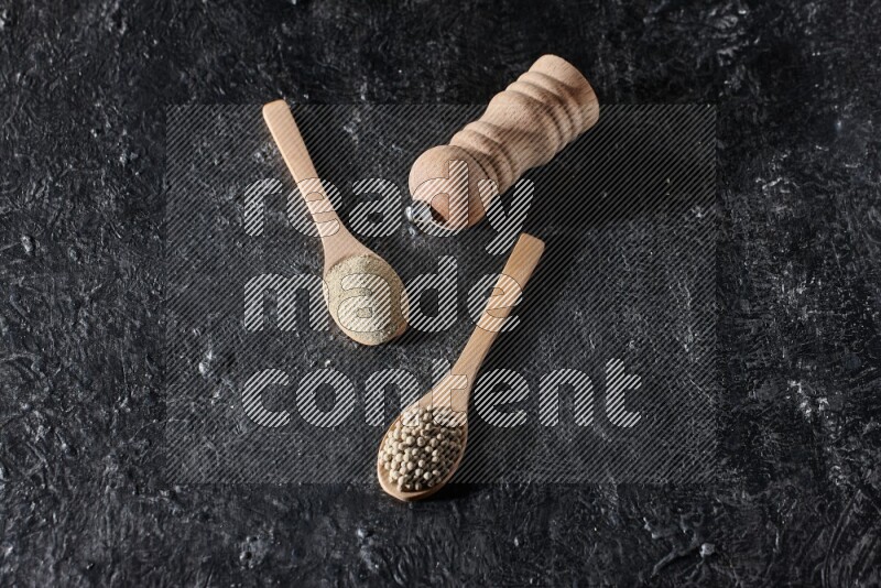 2 wooden spoons one full of white pepper powder and the other with pepper beads and a wooden pepper mill on textured black flooring