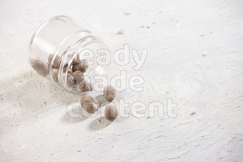 A glass jar full of nutmeg flipped and the seeds spread out on a textured white flooring