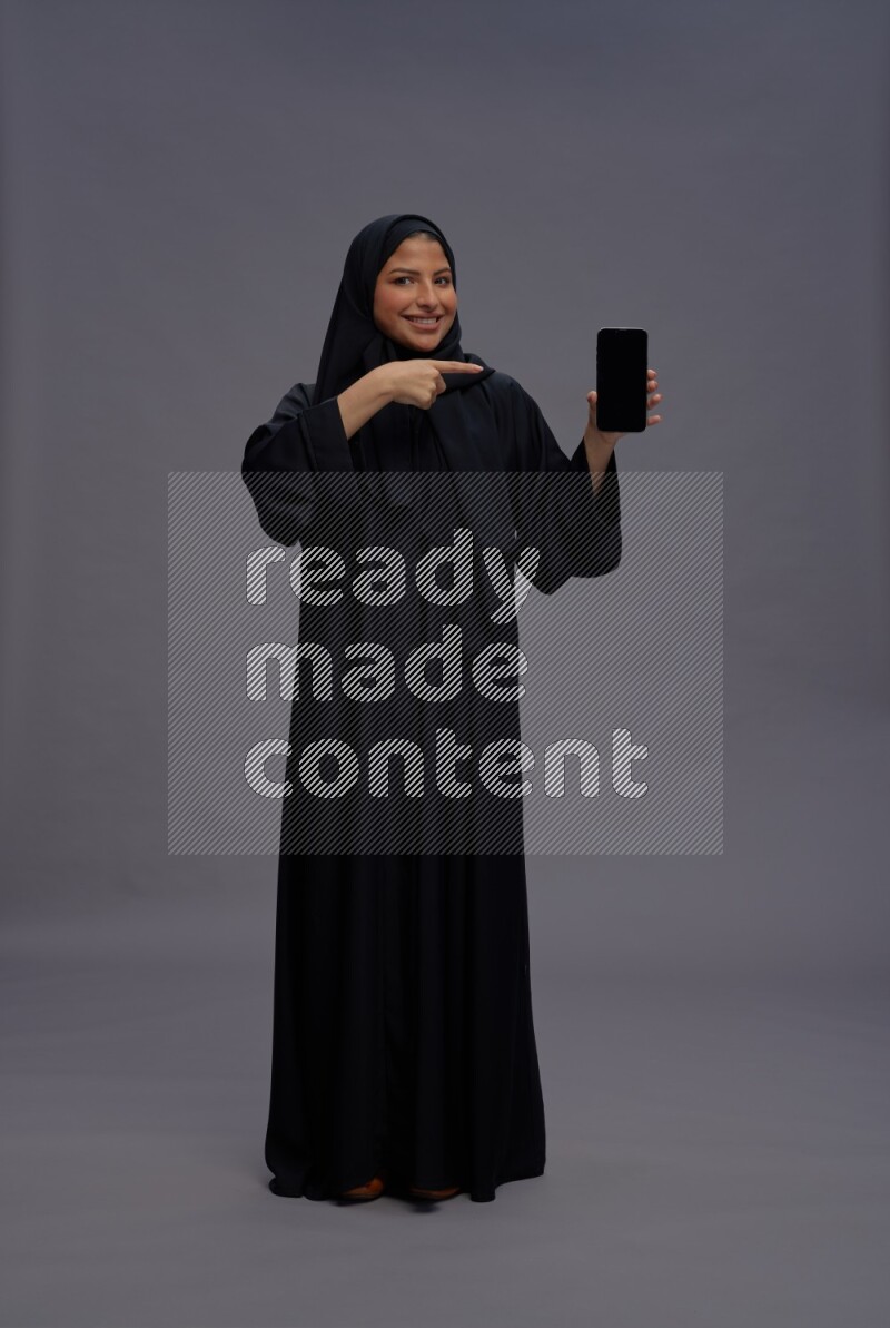 Saudi woman wearing Abaya standing showing phone to camera on gray background