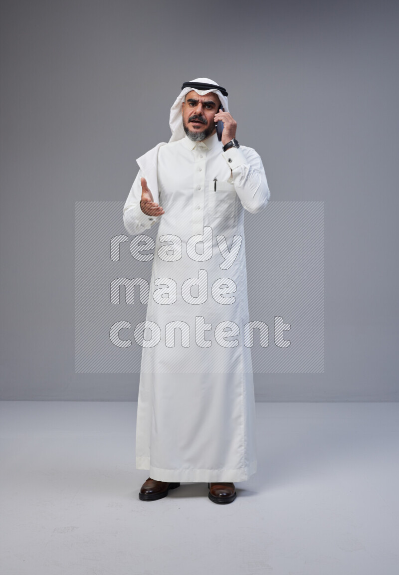 Saudi man Wearing Thob and white Shomag standing talking on phone on Gray background