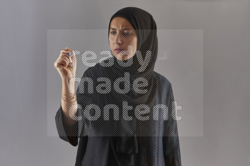 Woman in black abaya and hijab holding a marker pen to write on a transparent board with different reactions and expressions on grey background