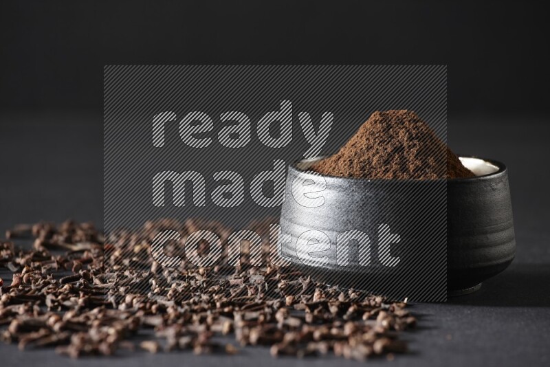 A black pottery bowl full of cloves powder and cloves whole grains on a black flooring