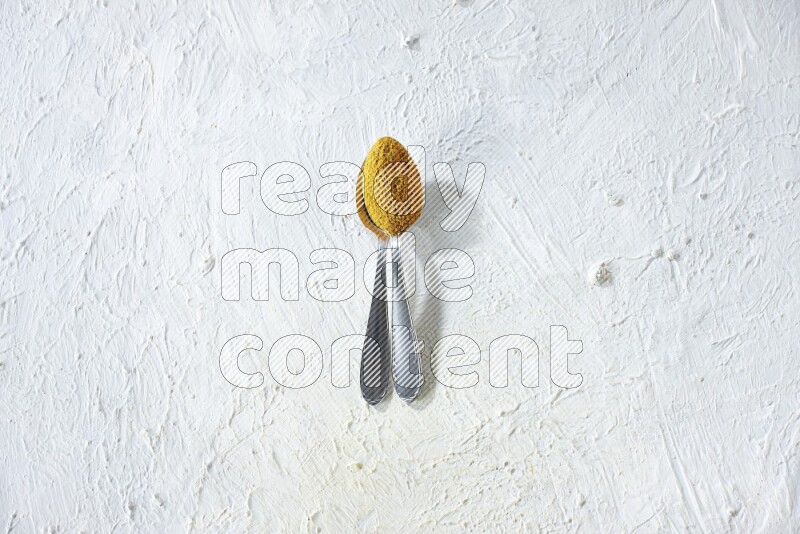 2 metal spoons full of turmeric powder on a textured white background