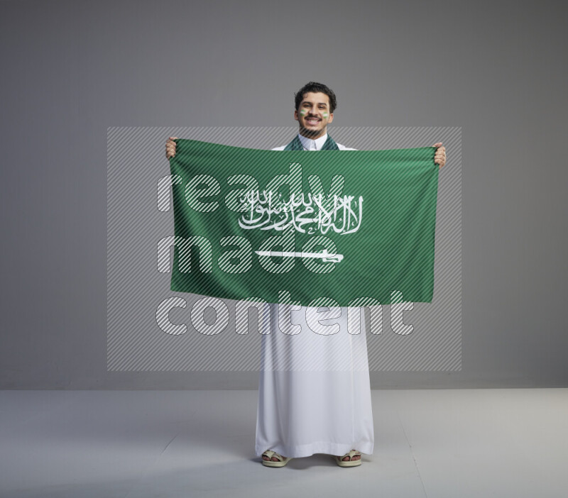 A Saudi man standing wearing thob with face painting wrapping Saudi flag scarf and holding big Saudi flag on gray background