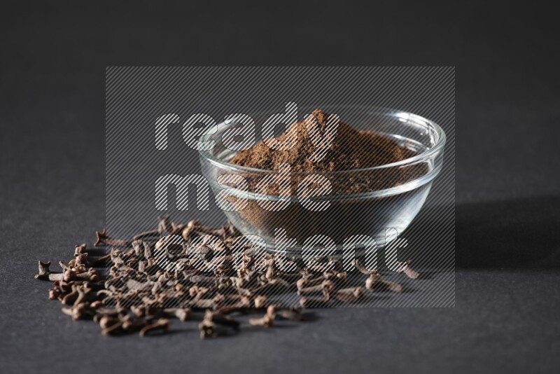 A glass bowl full of cloves powder with cloves grains spread on black flooring