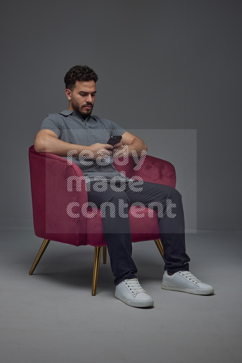 A man wearing casual and using his phone while sitting on a burgundy chair eye level on a gray background