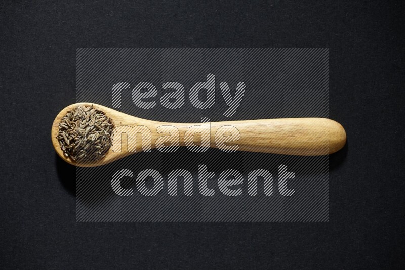 A wooden spoon full of cumin seeds on black flooring