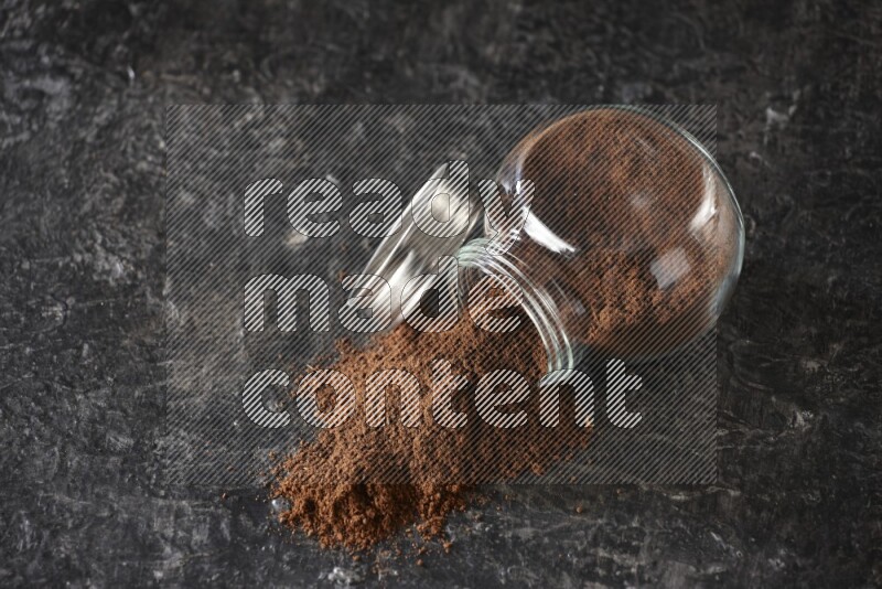 A flipped glass spice jar full of cloves powder and powder came out of it on textured black flooring