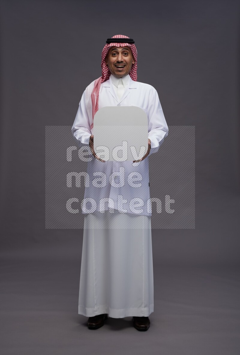 Saudi man wearing thob with lab coat and shomag with pocket employee badge standing holding social media sign on gray background