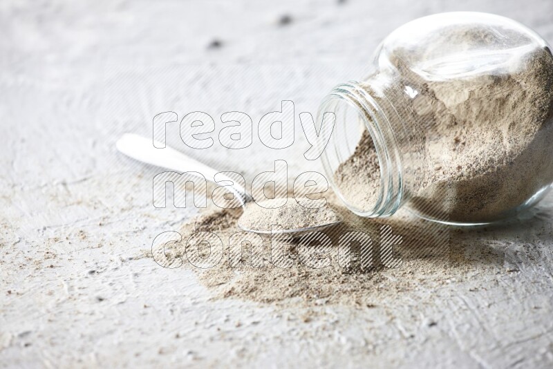 A flipped herbal glass jar and metal spoon full of white pepper powder with spilled powder on textured white flooring
